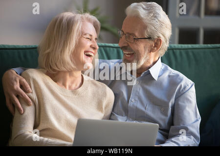 Happy older spouses couple laughing embracing using laptop at home Stock Photo