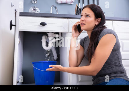 Worried Woman Calling Plumber While Collecting Water Leaking From Sink Using Utensil Stock Photo