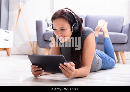 Happy Young Woman Lying On Floor Enjoying Music On Headphone Using Digital Tablet Stock Photo