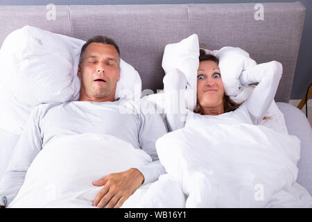 Mature Woman Covering Her Ears With Cushion While Man Snoring In Bed Stock Photo