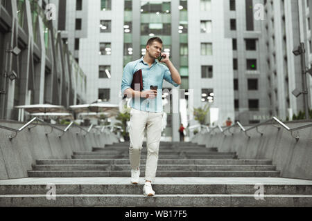 young handsome office worker talking on smartphone holding laptop and cup of coffee walking down the stairs at business center Stock Photo