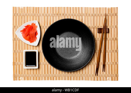 Empty round black plate with chopsticks for sushi and soy sauce, ginger on yellow bamboo mat background. Top view with copy space for you design. Stock Photo