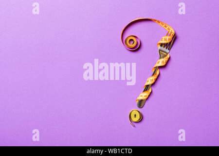Top view of fork and measure tape in form of question mark on purple background. Overeating concept and copy space. Stock Photo