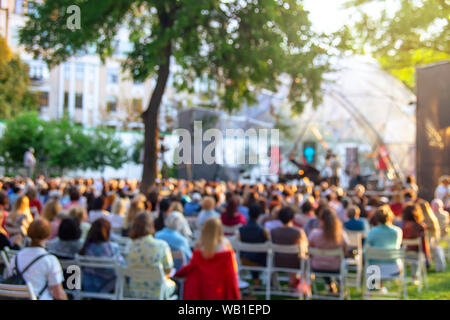 Summer street concert on an open stage with spectators sitting on chairs on the grass under a tree. Blurred image, background Stock Photo