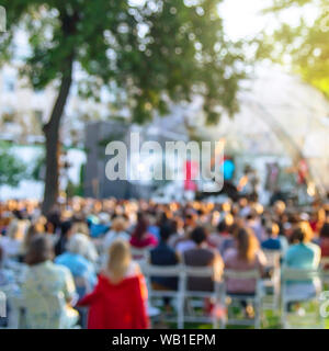 Summer street concert on an open stage with spectators sitting on chairs on the grass under a tree. Blurred image, background Stock Photo