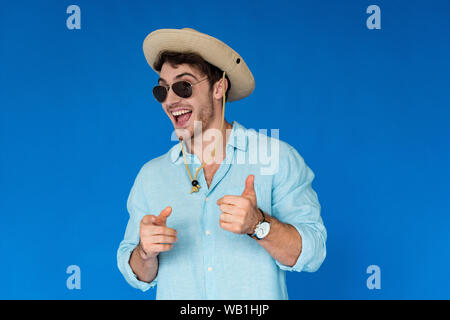 smiling traveler in safari hat showing thumb up and pointing with finger at camera isolated on blue Stock Photo