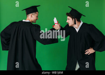 two students in academic caps holding diplomas and looking at each other isolated on green Stock Photo