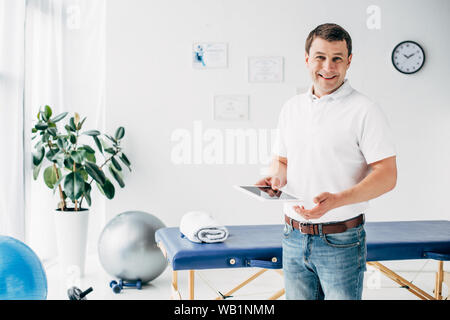 smiling chiropractor holding Digital Tablet in hospital and looking at camera Stock Photo