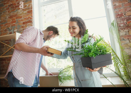 New life. Young couple moved to a new house or apartment. Look happy and confident. Family, moving, relations, first home concept. Unpacking boxes with their plants, books, put things on shelves. Stock Photo