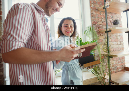 New life. Young couple moved to a new house or apartment. Look happy and confident. Family, moving, relations, first home concept. Unpacking boxes with their plants, books, put things on shelves. Stock Photo