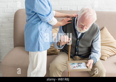 Nurse putting hands on upset and grey haired man with photo frame Stock Photo