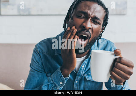 young african american man holding coffee cup while suffering from toothache