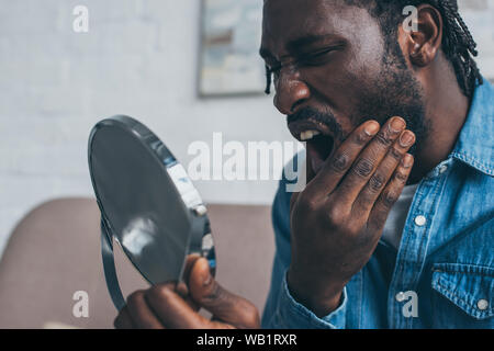 upset african american man looking at mirror while suffering from tooth pain Stock Photo