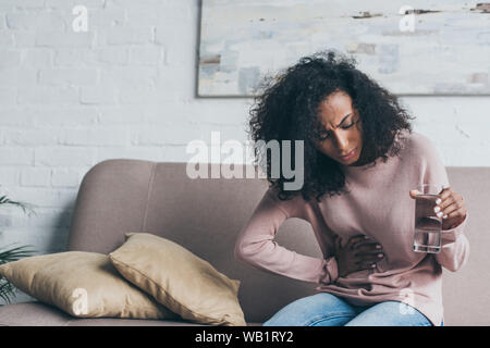 unhappy african american woman holding glass of water while sitting on sofa and suffering from stomach pain Stock Photo