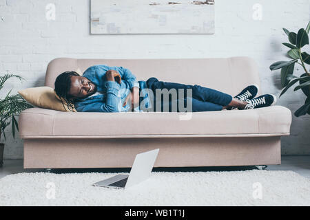 young african american man suffering from stomach pain while lying on sofa near laptop on floor Stock Photo