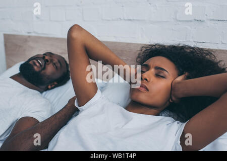 exhausted african american woman covering ears with hands while lying near snoring husband Stock Photo