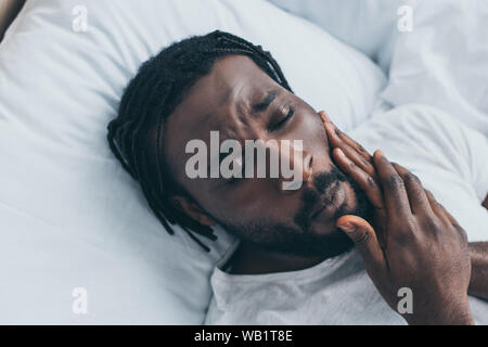 unhappy african american man suffering from jaw pain while lying in bed