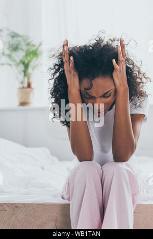 unhappy african american woman holding hands near head while suffering from headache in bedroom Stock Photo