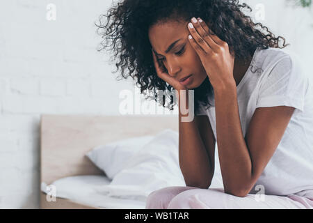 pretty african american woman holding hands near head while suffering from headache in bedroom Stock Photo