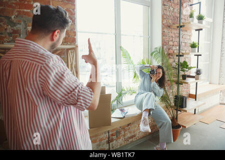 New life. Young couple moved to a new house or apartment. Look happy and confident. Family, moving, relations, first home concept. Unpacking boxes with their plants, books, put things on shelves. Stock Photo