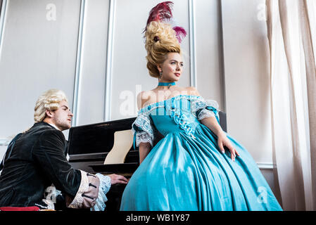 attractive victorian woman in blue dress standing near man in wig playing piano Stock Photo