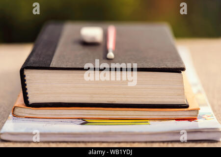 A stack of books & notebooks with pencil and eraser on a wooden desk at outdoor park. Background concept for education loan, scholarship, teachers day Stock Photo
