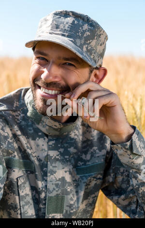 happy soldier in military uniform and cap holding wheat near lips Stock Photo