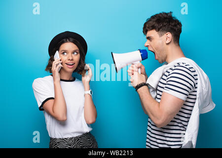 irritated man quarreling in loudspeaker at girlfriend talking on smartphone on blue background Stock Photo