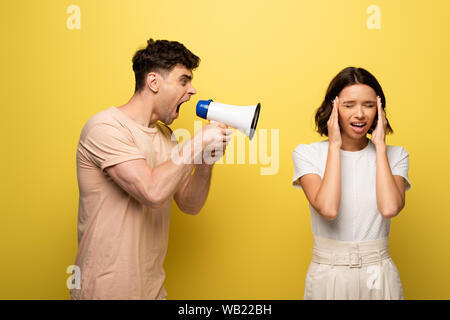 angry young man quarreling in loudspeaker at girfriend covering ears with hands on yellow background Stock Photo