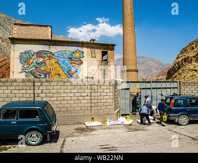 A Soviet-era mosaic depicting a miner on a factory wall in Zeravshan II, Tajikistan, recalls the region's former mining industry, men unloading sacks of food from a truck in front of it. Stock Photo