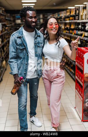 happy african american man holding bottle near asian girl in sunglasses pointing with finger in supermarket Stock Photo