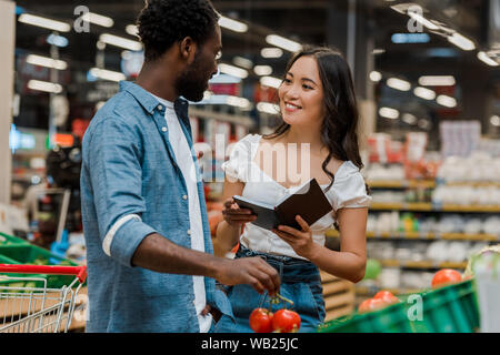 african american man holding fresh tomatoes and looking at cheerful asian woman with notebook Stock Photo