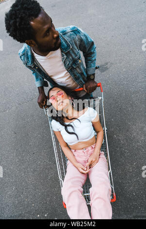overhead view of handsome african american man standing near asian girl sitting in shopping trolley outside Stock Photo