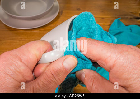 Close overhead shot of a man’s hands using a tea towel to dry a wet cup, next to a dish and plate, on a pine kitchen worktop. Stock Photo