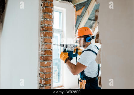 selective focus of workman in uniform and yellow gloves using hammer drill on brick wall Stock Photo