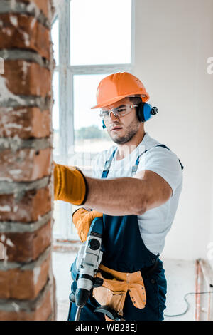 selective focus of workman in uniform and yellow gloves holding hammer drill and looking at brick wall Stock Photo
