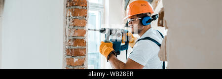 panoramic shot of handyman in uniform and yellow gloves using hammer drill Stock Photo
