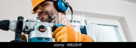 panoramic shot of handyman in helmet using hammer drill Stock Photo