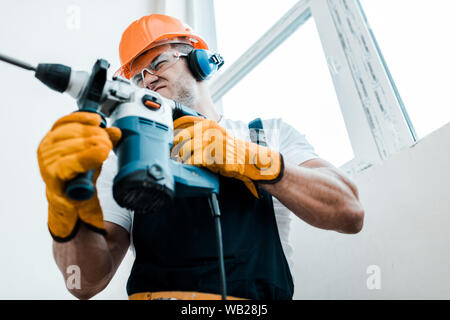 low angle view of handyman in helmet and yellow gloves using hammer drill Stock Photo