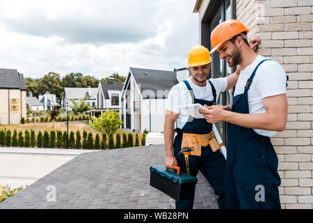 happy handyman holding toolbox near cheerful coworker using digital tablet Stock Photo