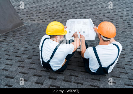 overhead view of architects in helmets sitting on roof with blueprint Stock Photo
