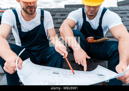 cropped view of architect in helmet holding pencil near blueprint and coworker pointing with finger Stock Photo