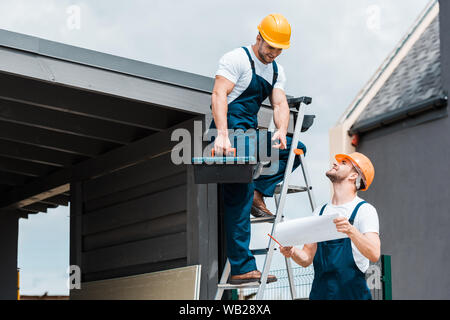 cheerful architect pointing with finger at paper near happy coworker in helmet Stock Photo