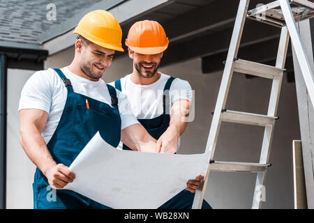 happy architects in helmets looking at paper and smiling near ladder Stock Photo