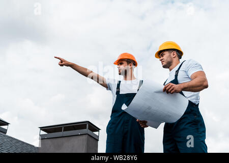 low angle view of handsome architect pointing with finger near coworker in helmet holding paper Stock Photo