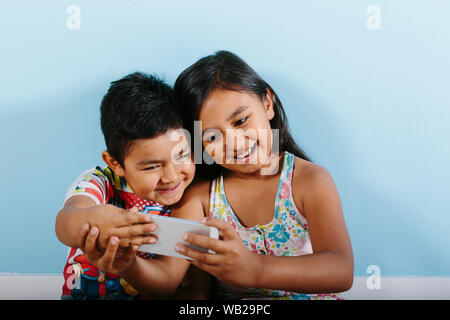 Two siblings, a 9-year old girl and a 5-year old boy are taking a selfie with a smartphone. They're smiling. They have hispanic features. They're lean Stock Photo