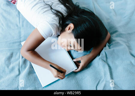 A 9-year old girl is lying on a blanket on bed. She's writing on a notebook. She has long dark hair. She has hispanic ethnicity features. Stock Photo