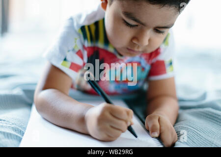 A 5-year old boy is lying on a blanket on bed. He's writing on his notebook. He has hispanic ethnicity features. He reveals concentration and attentio Stock Photo