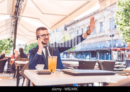 Handsome businessman in glasses waving to someone while talking on the phone during coffee break in a cafe. Stock Photo