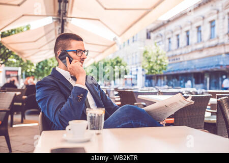 Young businessman in glasses sitting at cafe, reading newspapers and talking on mobile phone. Stock Photo